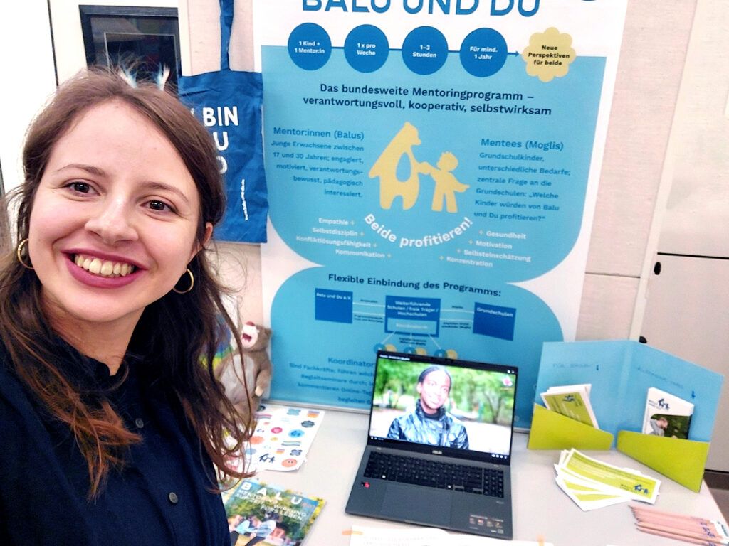 Foto: Regionalmanagerin Elena Klein vor einem Stand, auf dem Infomaterial zu Balu und Du gezeigt werden.