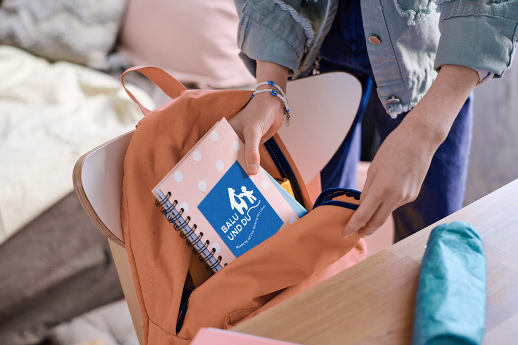 Foto von den Händen einer Person, die ein Ringbuch mit der Aufschrift "Balu und Du" aus einem Rucksack nimmt
