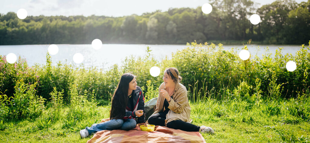 Foto: Zwei junge Frau sitzen an einem See auf einer Decke, vor ihnen steht eine Schale mit etwas zu Essen.