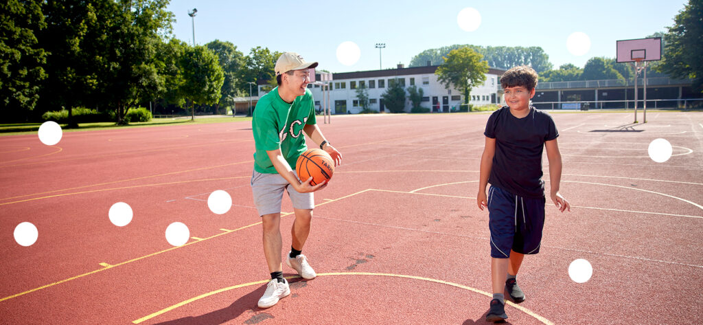 Foto, auf dem ein junger Erwachsener und ein Junge im Freien Basketball spielen.